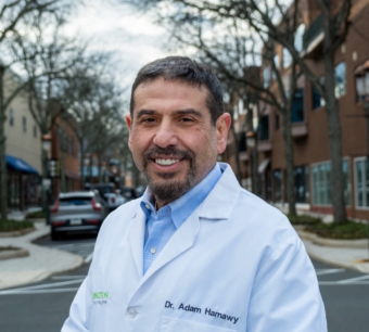 Adam Hamawy Portrait photo of Adam Hamawy in a medical jacket, with a neighborhood in the background.